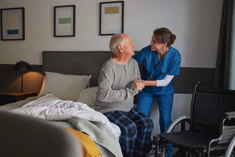 Two older adults sit together at a dining table, talking with a staff member in a bright, home-like common room.