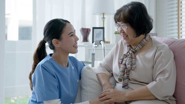 A senior resident sitting in a sunny lounge with a small dog on their lap and a window view of a garden outside.