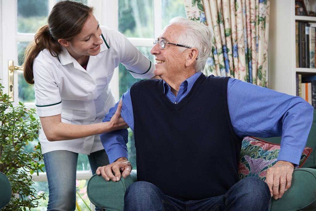 Family members sit with an older resident in a bright assisted living room, talking beside family photographs.