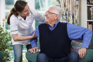 Family members sit with an older resident in a bright assisted living room, talking beside family photographs.